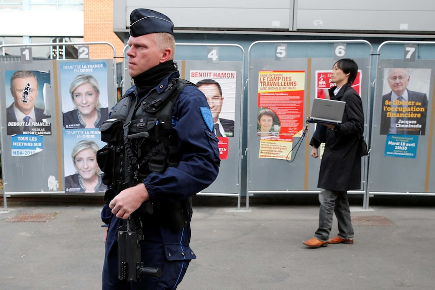 A police officer with gun, a person walks behind him, posters of the candidates in the background.