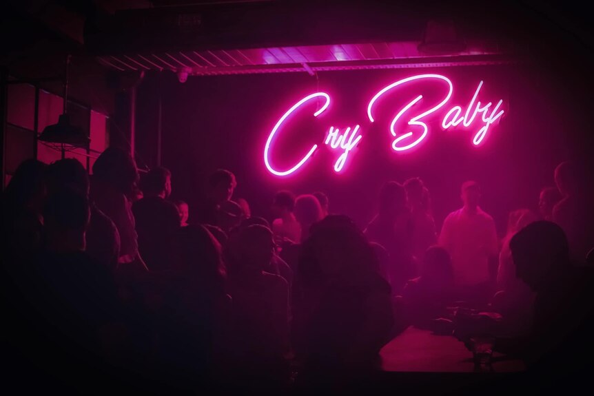 A pink neon sign saying Cry Baby in a dark nightclub 