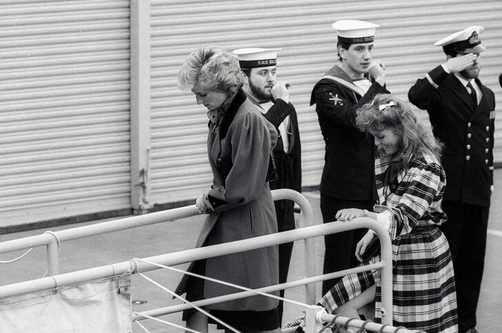 Diana looks down as she boards ship next to Sarah Ferguson in check coat, who holds onto railing. Three sailors are seen behind.