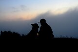 A man and a dog sit together in silhouette, in front of a beautiful sunset.