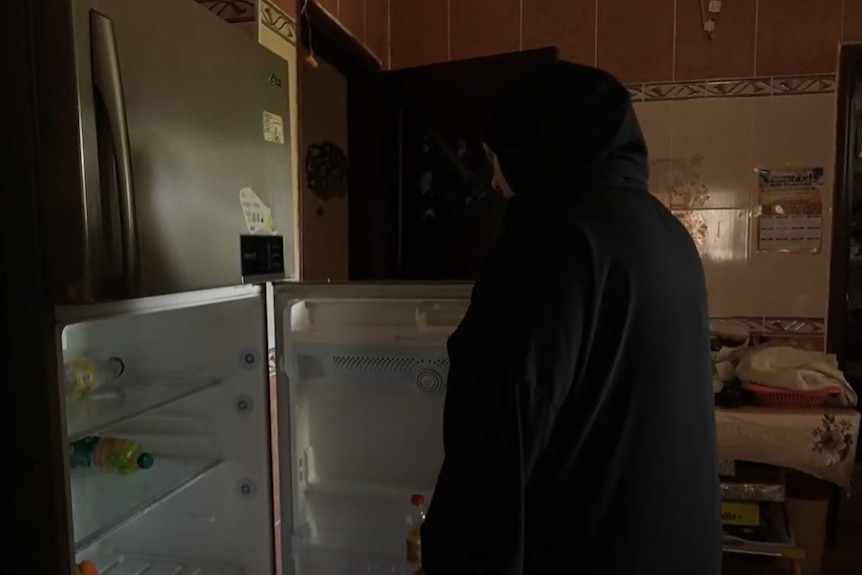 Woman in hijab opens a fridge which only has a few bottles and no electricity. 
