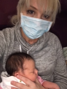 A woman wearing a mask and a grey jumper holds a sleeping newborn baby against her chest
