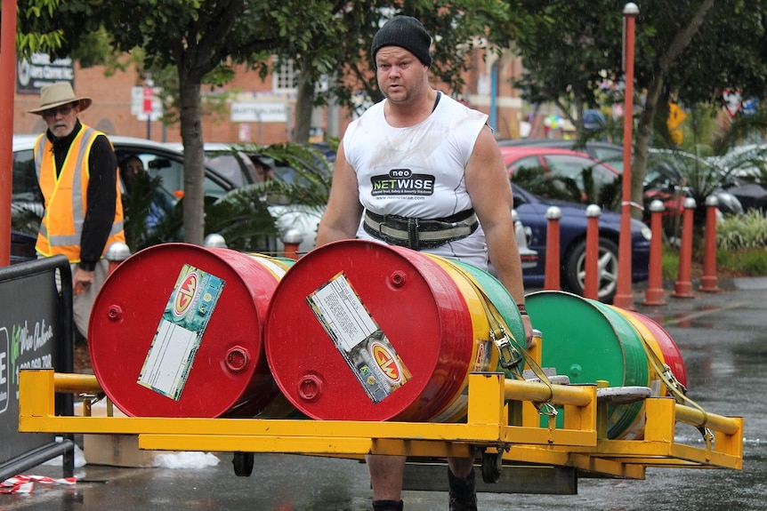 A man in a white singlet carrying a frame with four 44 gallon drums