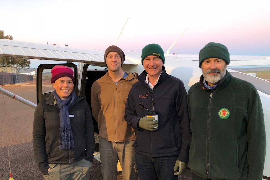A woman and three men, all wearing beanies, stand smiling in front of a small aeroplane.