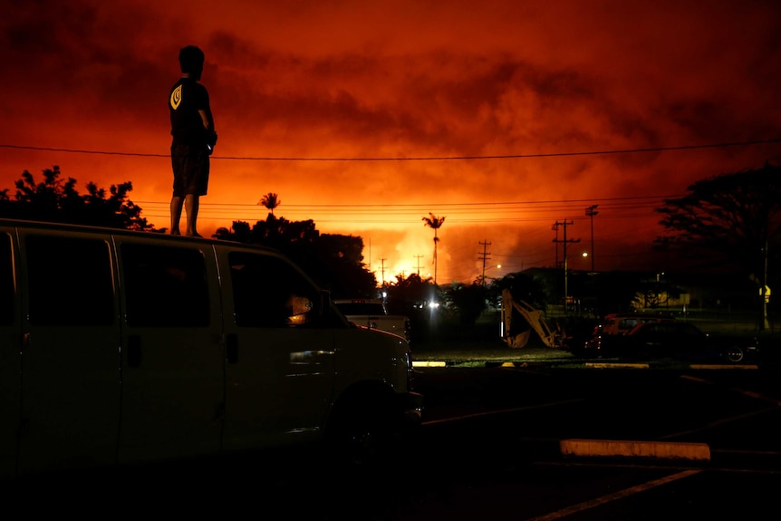 A man stands on a car and look out towards a bright orange sky created by an erupting volcano.