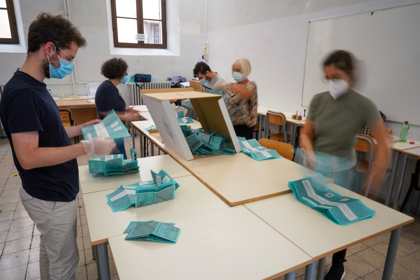 Several people wearing masks and gloves stand around tables counting blue ballots.