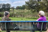 Two women sit on opposite ends of a park bench as they talk to each other.