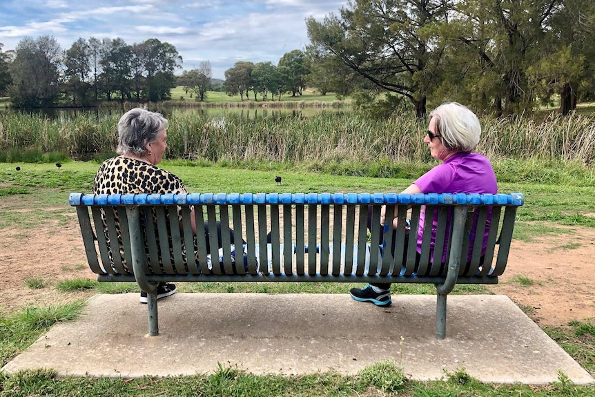 Two women sit on opposite ends of a park bench as they talk to each other.