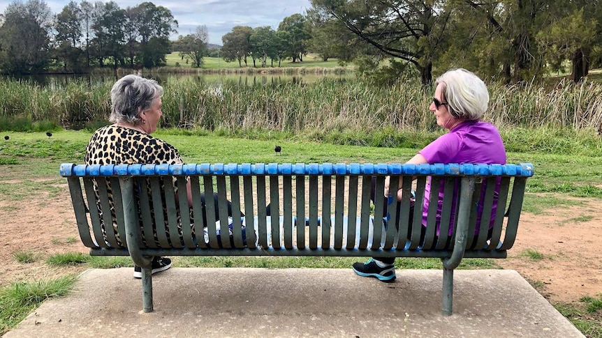 Two women sit on opposite ends of a park bench as they talk to each other.