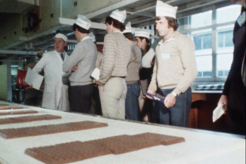 A grainy photo of a group of men wearing white paper hats looking a table with uncovred chocolate blocks