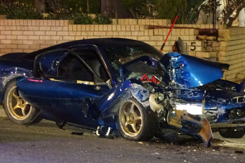 A blue car lies mangled on the road in front of a brick wall.