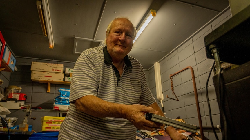 An elderly man leaning on a cane holds a video cassette tape and looks directly into the camera.