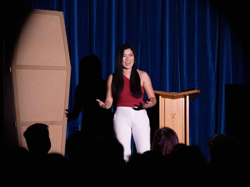 Annie Louey performing stand up in a red top and white pants, on stage with blue curtains as a backdrop