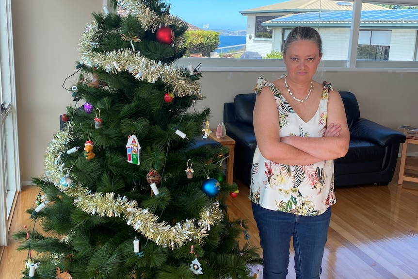 A woman stands next to a Christmas tree with her arms folded across her chest.