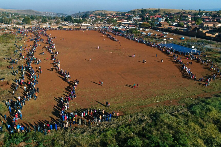 A huge queue of people stretches across a dirt clearing with houses seen on hills in the background.