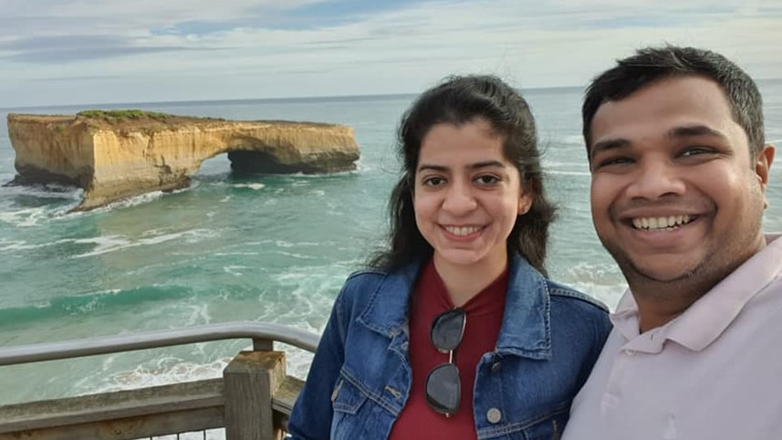 A smiling woman and man take a selfie in front of one of the 12 Apostles, a large stone in water.