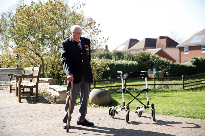 Tom Moore stands with a walking stick on a sunny day in his garden, with his walker nearby.