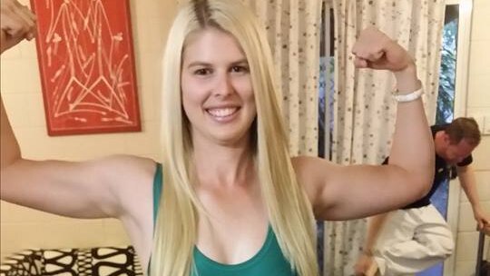 A young powerlifter flexes her muscles and smiles while wearing a green and gold bodysuit.