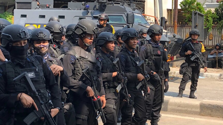 Heavily armoured Indonesian police officers stand on the street in front of a tank.