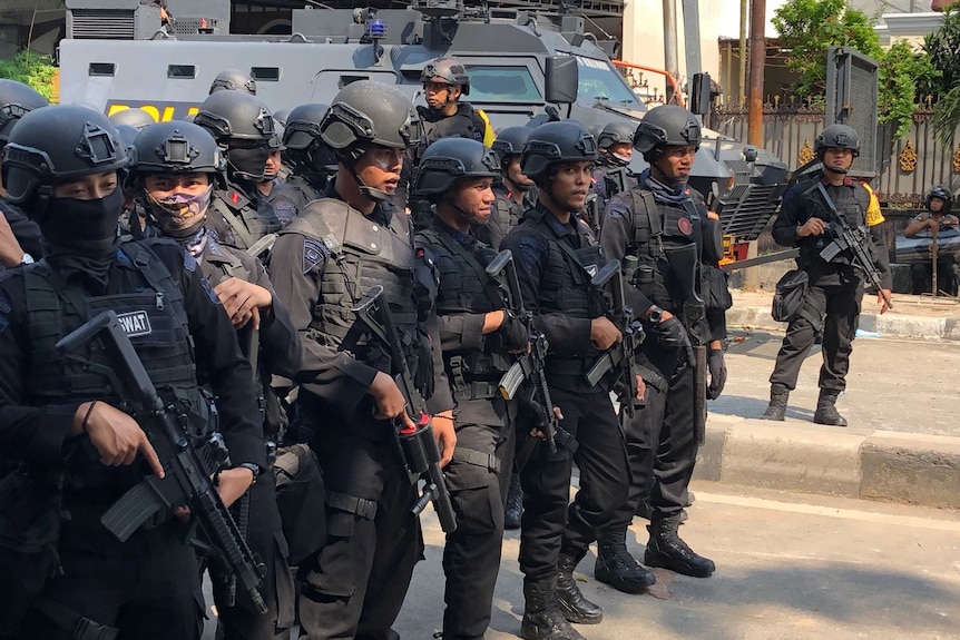 Heavily armoured Indonesian police officers stand on the street in front of a tank.