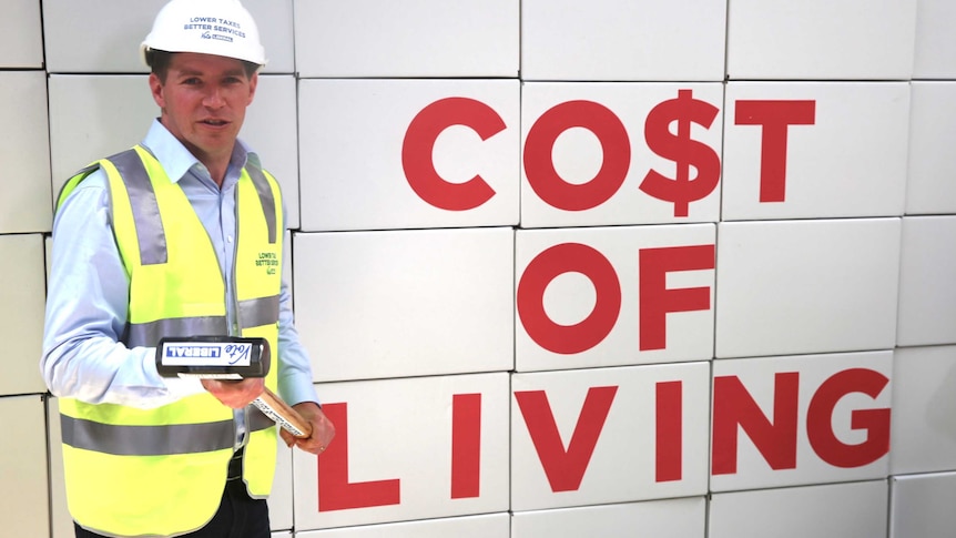 Alistair Coe holds a sledge hammer in front of boxes labelled "co$t of living".