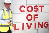 Alistair Coe holds a sledge hammer in front of boxes labelled "co$t of living".