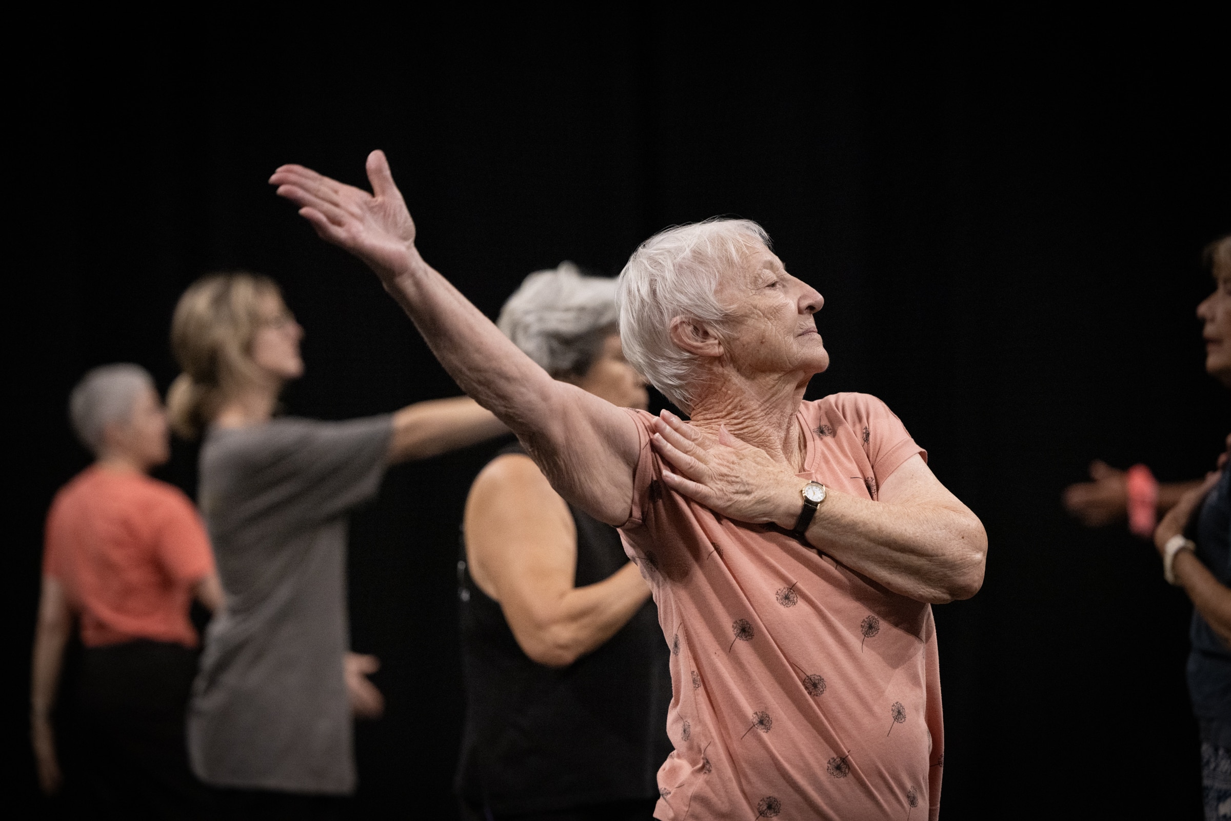 An older woman with short grey hair in a pink top stretches her arm backwards.
