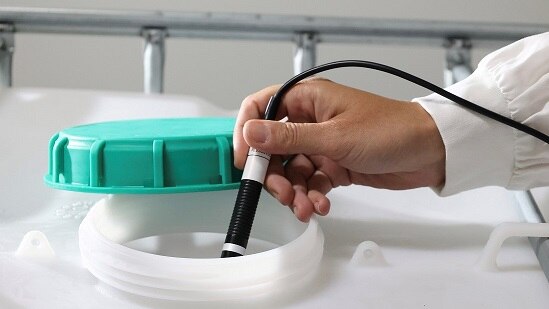 Hand in a white sleeve holds a black and white probe into a large plastic container with a green lid to the side