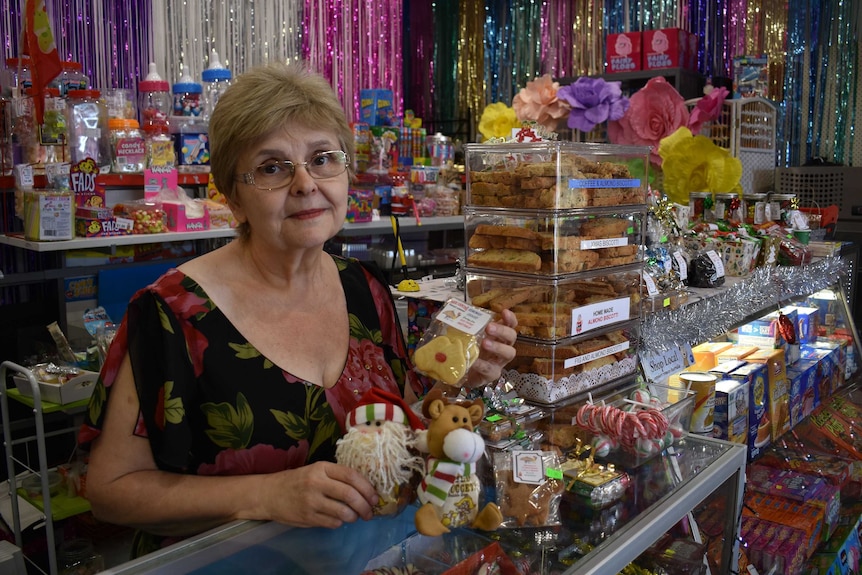 Silvana Placentino is behind the counter of a lolly shop holding cookies in her hand. She is wearing a floral shirt.