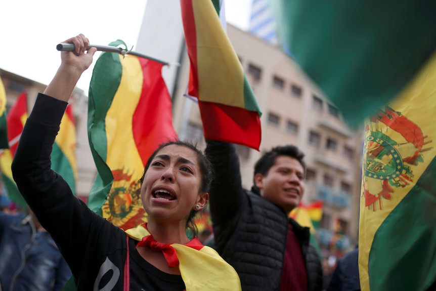 A woman with a pained expression, stands in a crowd of protesters waving Bolivian flags.