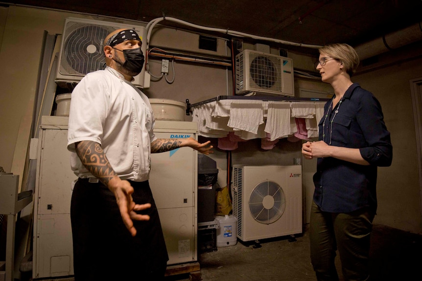 JC Miranda, dressed in a chef's jacket and apron, talks to Kate Cole in front of a ventilation system outside his restaurant.