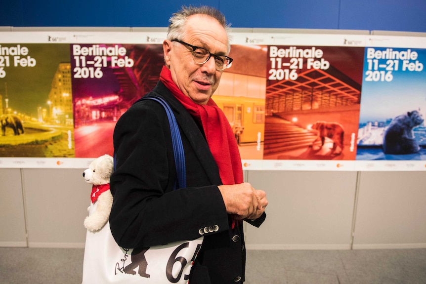 Dieter Kosslick with a shoulder bag containing a white teddy bear in front of coloured posters.