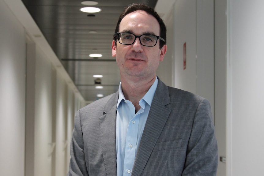 A photo of a man with glasses wearing a grey suit and blue shirt standing in corridor