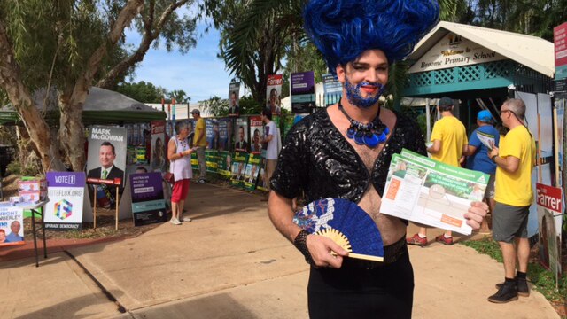 A drag queen dressed in black wearing a blue wig holding Greens how-to-vote cards.