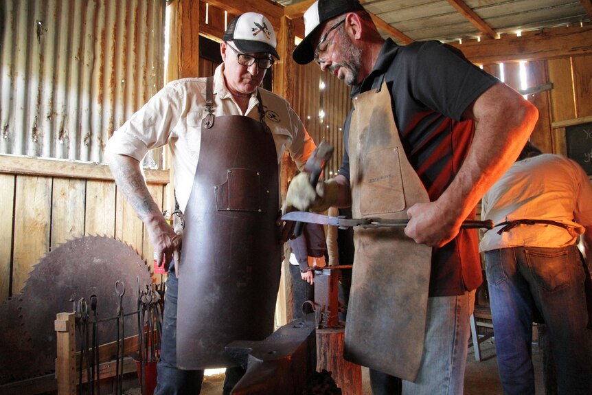 Two men inspecting a knife they are forging, both wear black and cream caps, leather aprons. Another man's back is towards them.