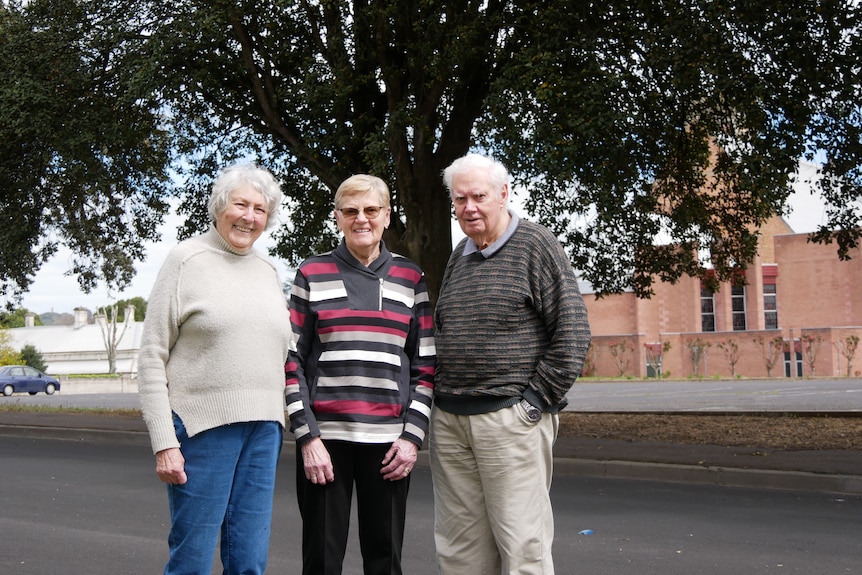 Three people standing in front of a large tree with a church in the background.