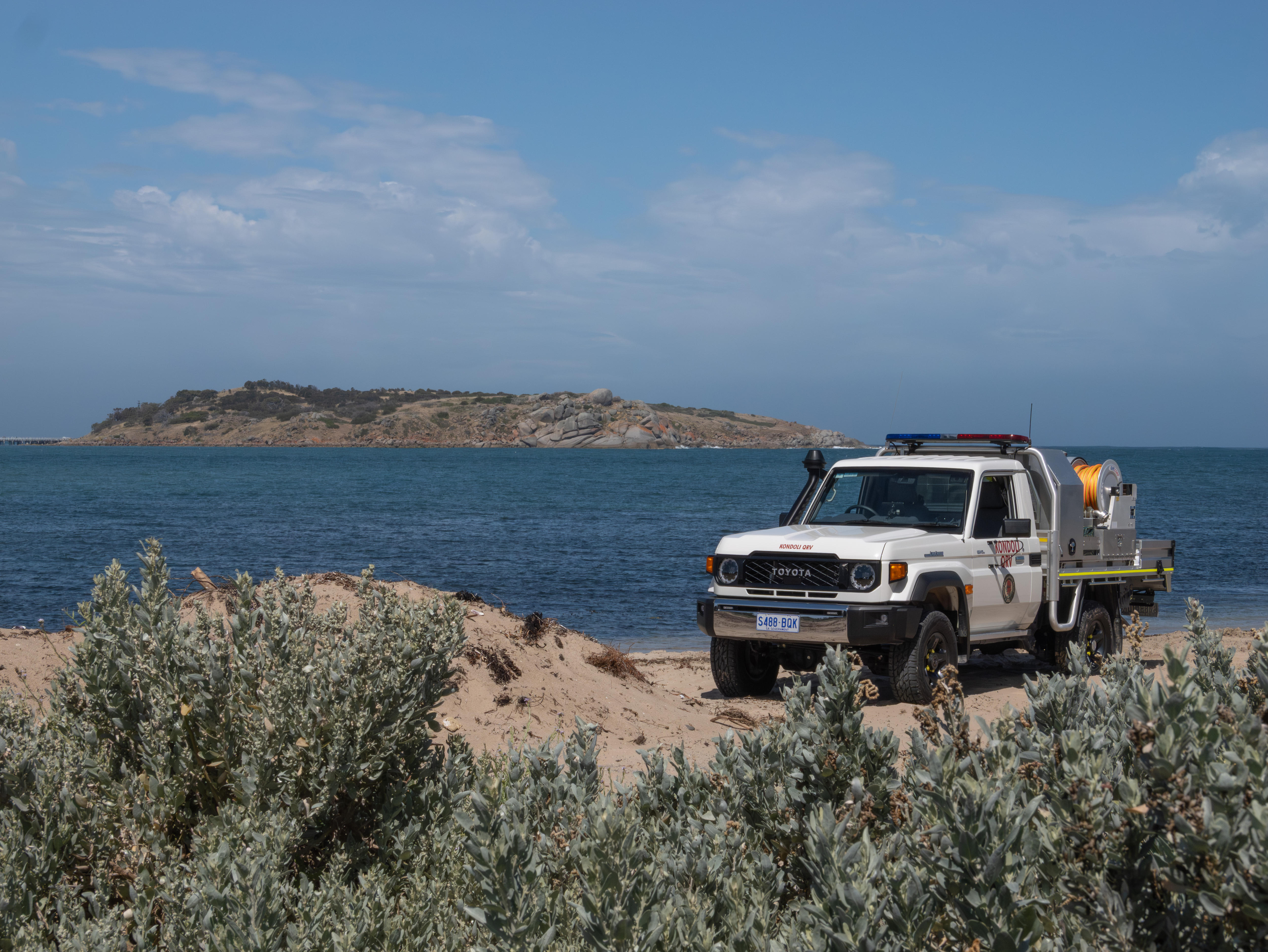 Un camión de bomberos blanco de cuatro ruedas sobre arena frente al mar. Arbustos nativos en primer plano. 