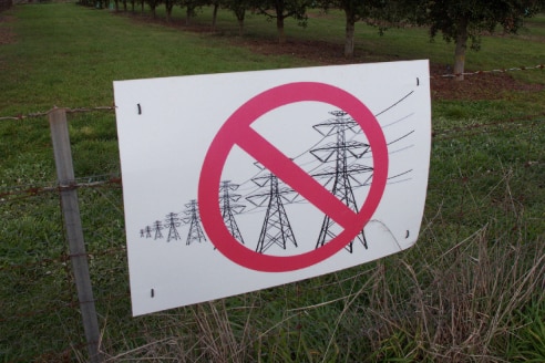 A sign depicting power lines with a cross over hangs on a farm gate.