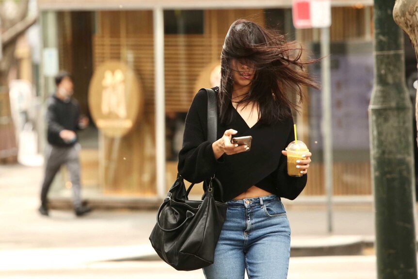 A woman wearing blue jeans and a black top has her hair blown into her face by wind as she walks.
