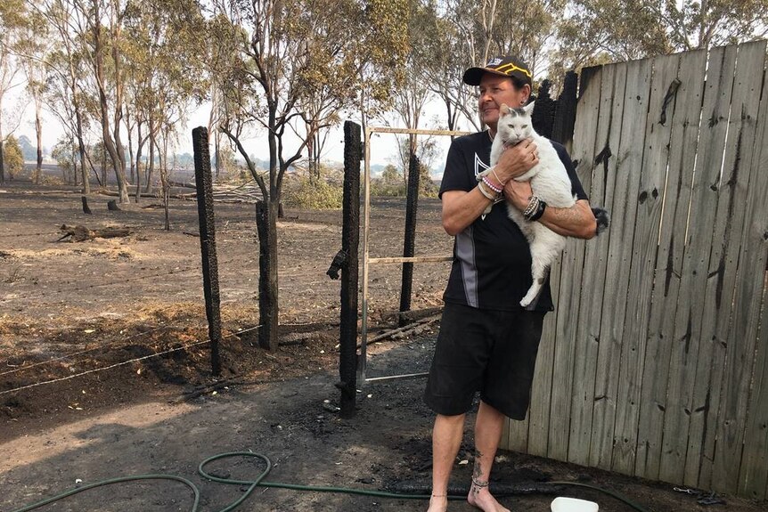 A man holds a white cat and looks out at the burnt property.