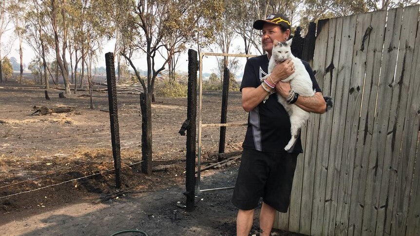 A man holds a white cat and looks out at the burnt property.