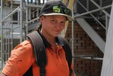 Emily Bailey smiles at the camera in a toolbelt and smudged shirt, with scaffolding emerging behind her on a worksite.