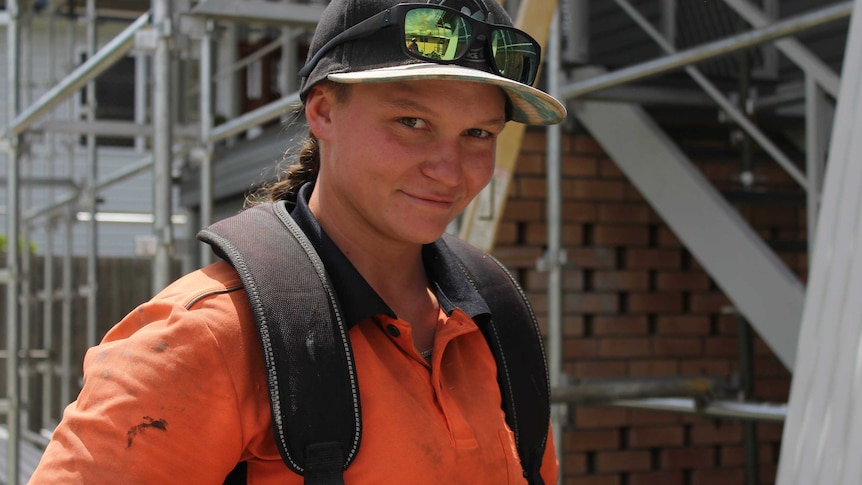 Emily Bailey smiles at the camera in a toolbelt and smudged shirt, with scaffolding emerging behind her on a worksite.
