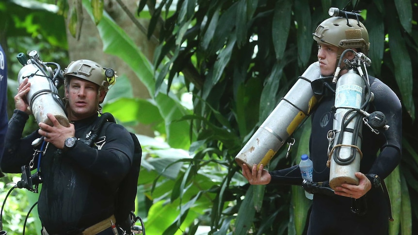 Two men in wetsuits and helmets with torches carry air tanks, standing against a lush rainforest background.