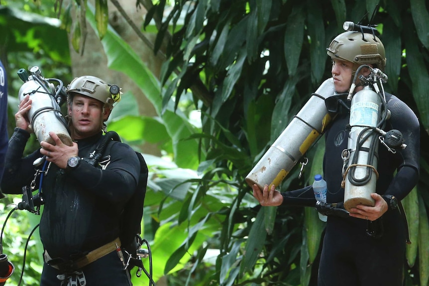 Two men in wetsuits and helmets with torches carry air tanks, standing against a lush rainforest background.