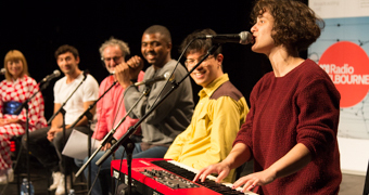 Five people with microphones watch as a woman plays keyboard and sings.