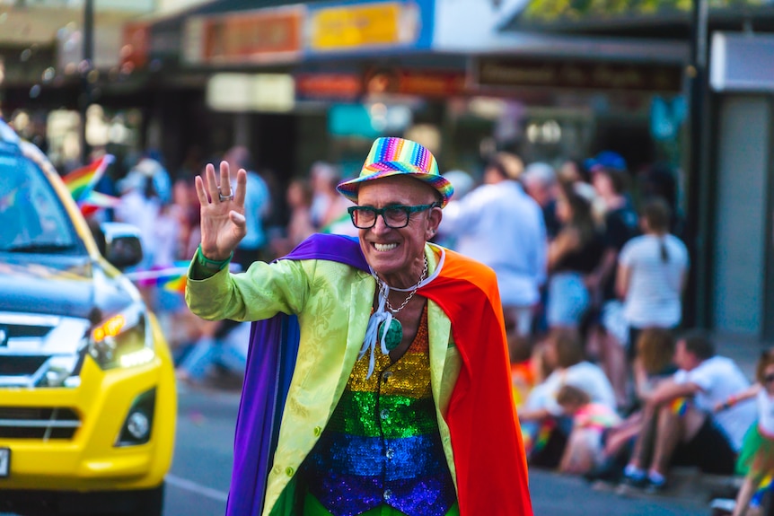 A man in glasses wearing rainbow attire waves to a crowd lining the street.