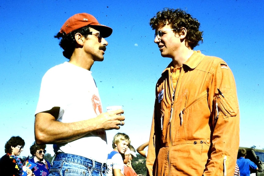 The winner of the 1988 balloon challenge wearing jeans and a white shirt talks with a man in an orange uniform with curly hair.