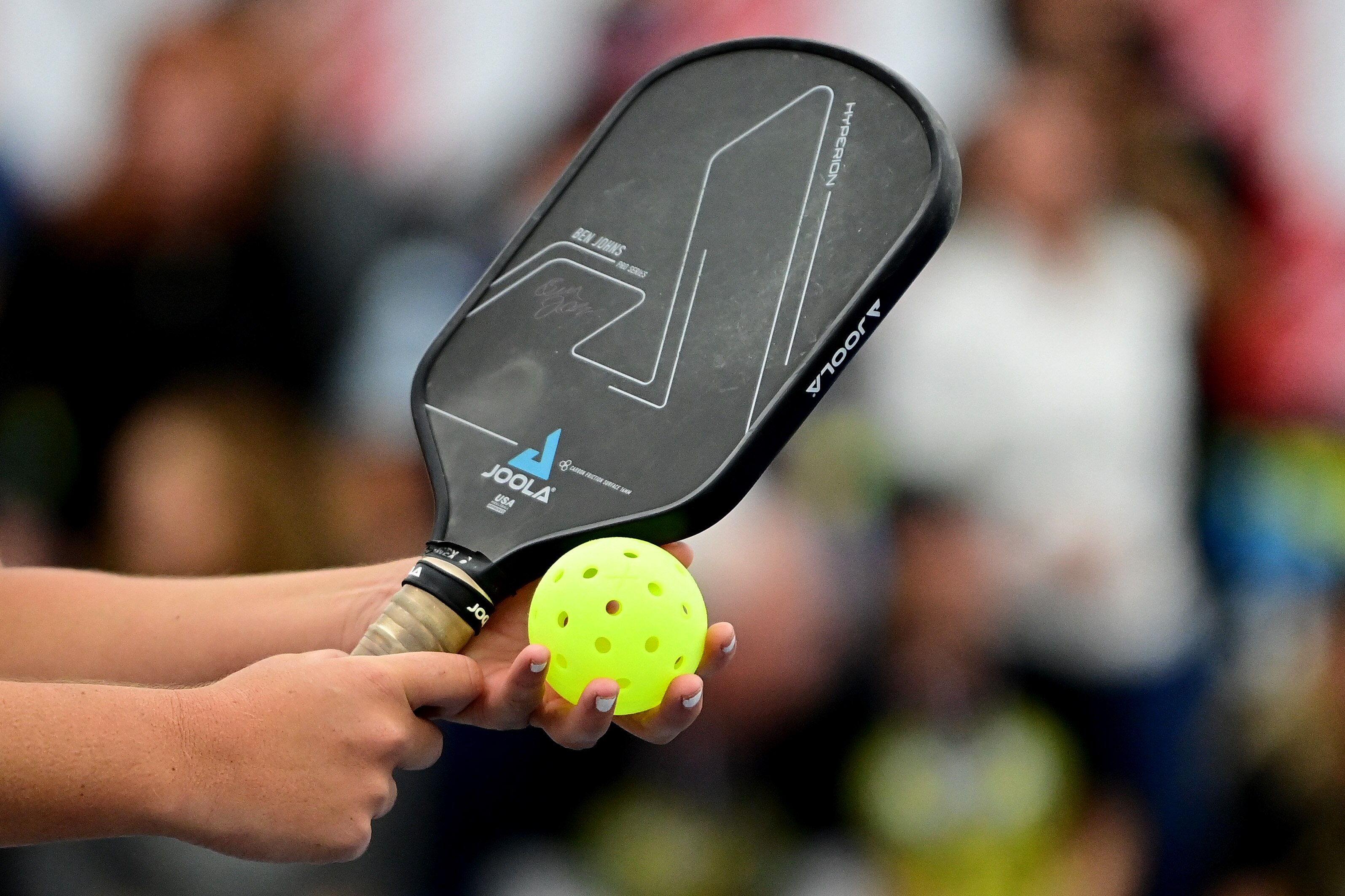 A pickleball paddle and ball are held by a player preparing to serve.