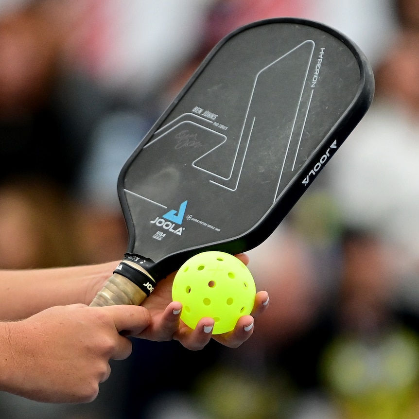 A pickleball paddle and ball are held by a player preparing to serve.